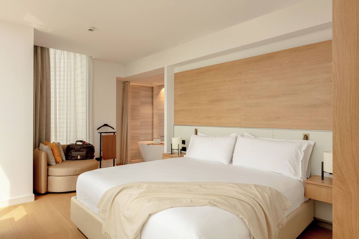 Una habitación de hotel moderna con una cama grande, una zona de estar con sofá y sillones, un cabecero de madera y un baño con bañera y espejo.