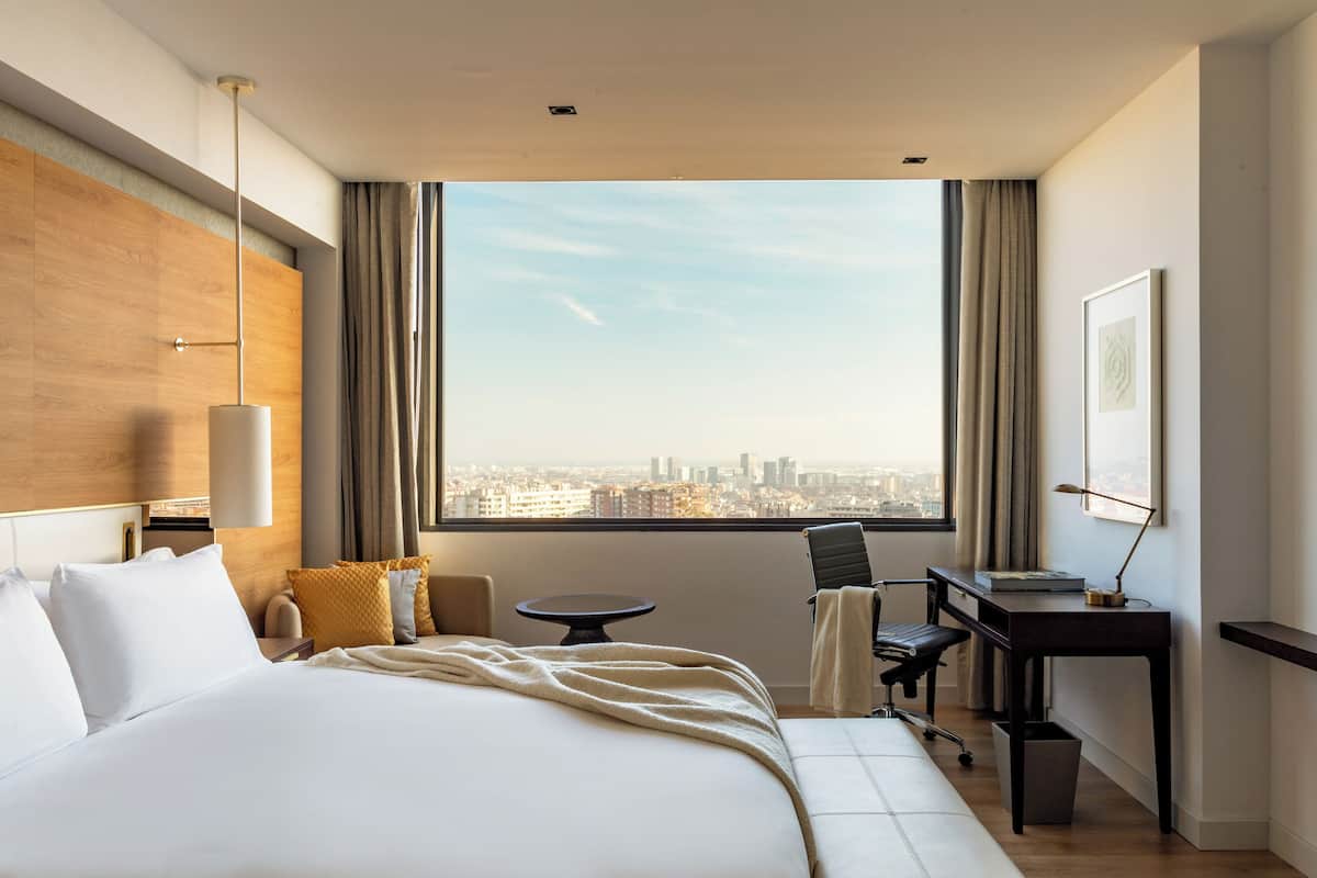 Habitación de hotel con una cama grande, un escritorio, una silla, una mesita y vistas a la ciudad.