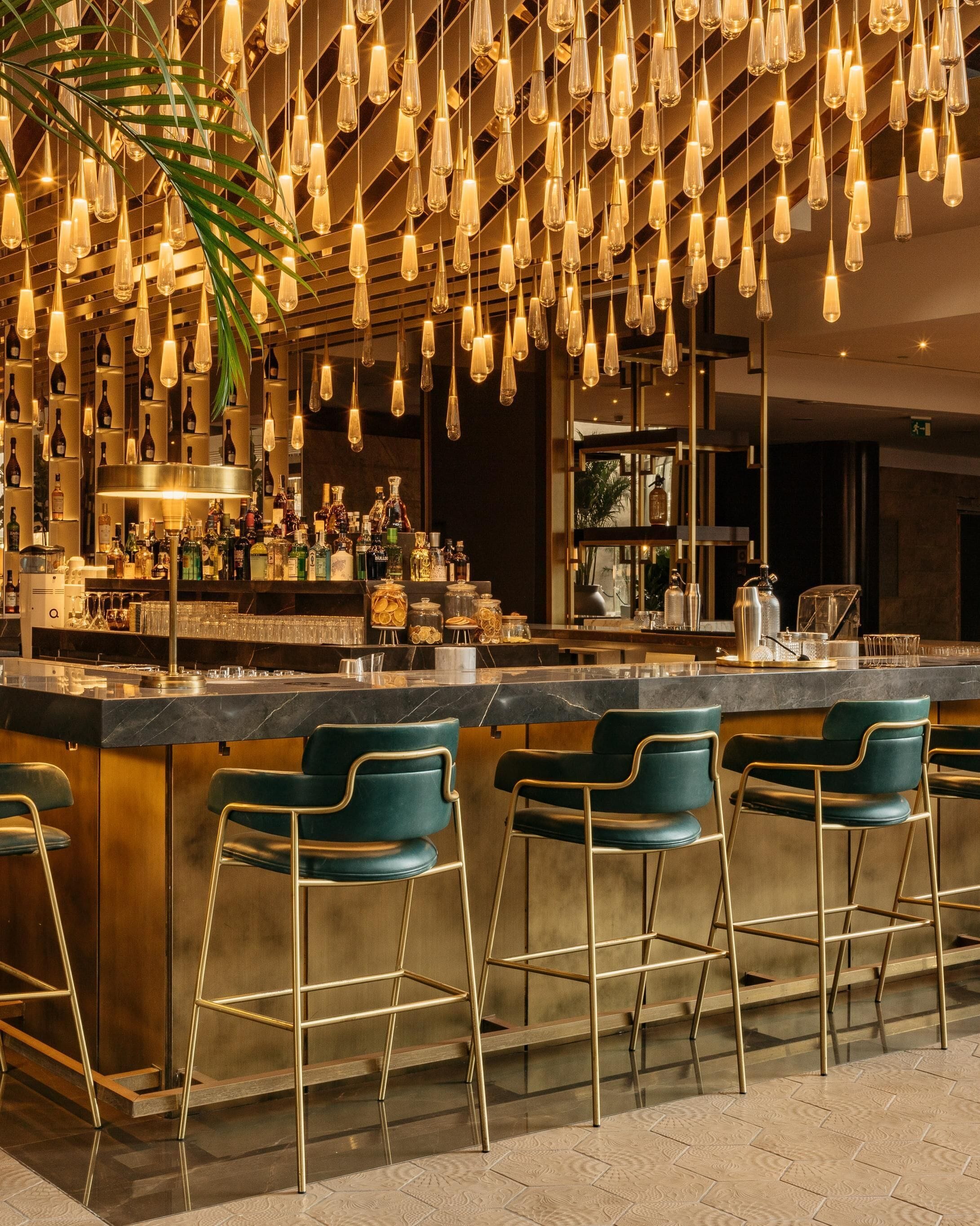 Zona de bar con lámparas colgantes, taburetes y una variedad de botellas y vasos en estantes.