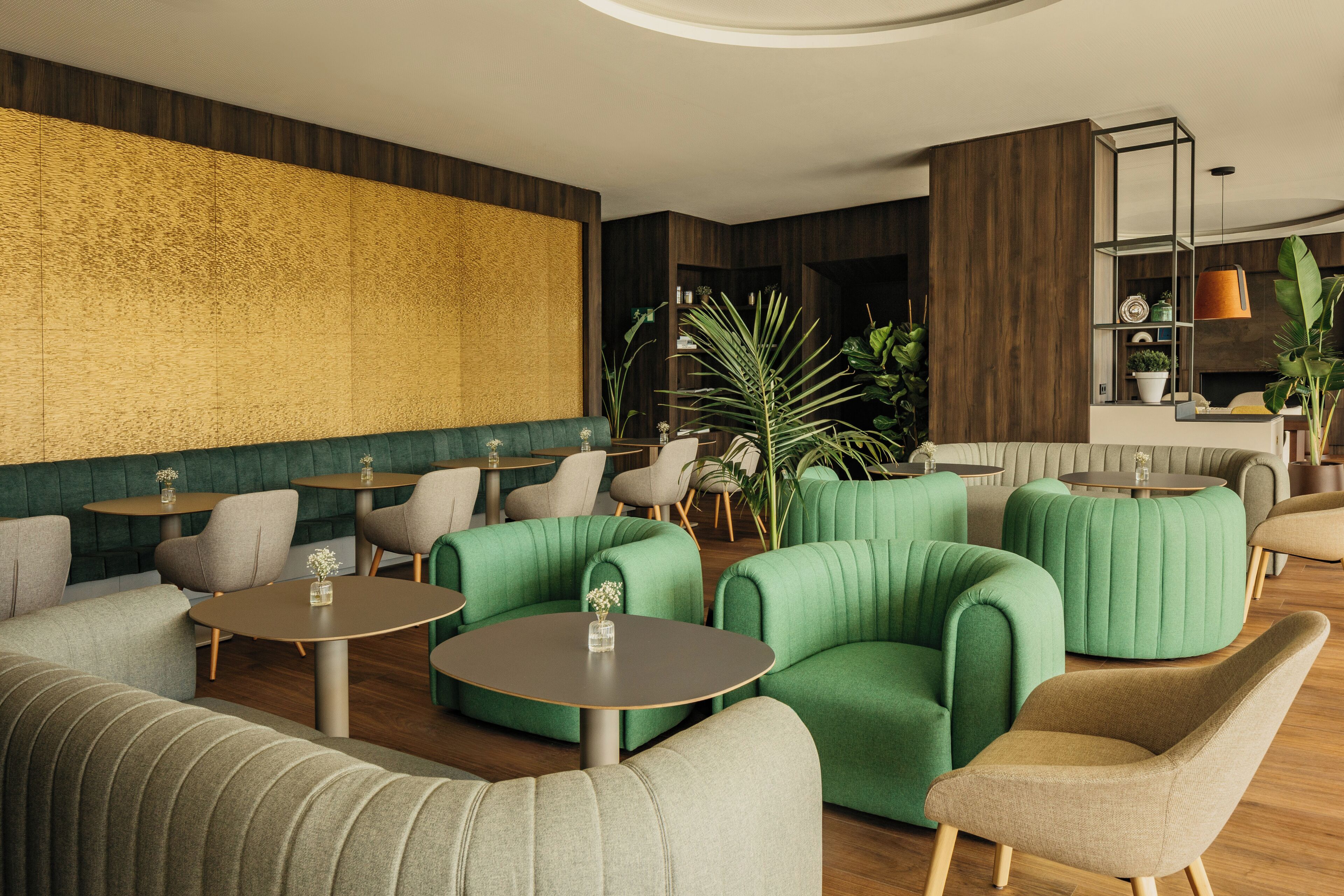 Un área de descanso moderna con asientos tapizados en verde, mesas redondas y una pared grande con textura.