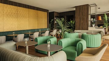Un área de descanso moderna con asientos tapizados en verde, mesas redondas y una pared grande con textura.