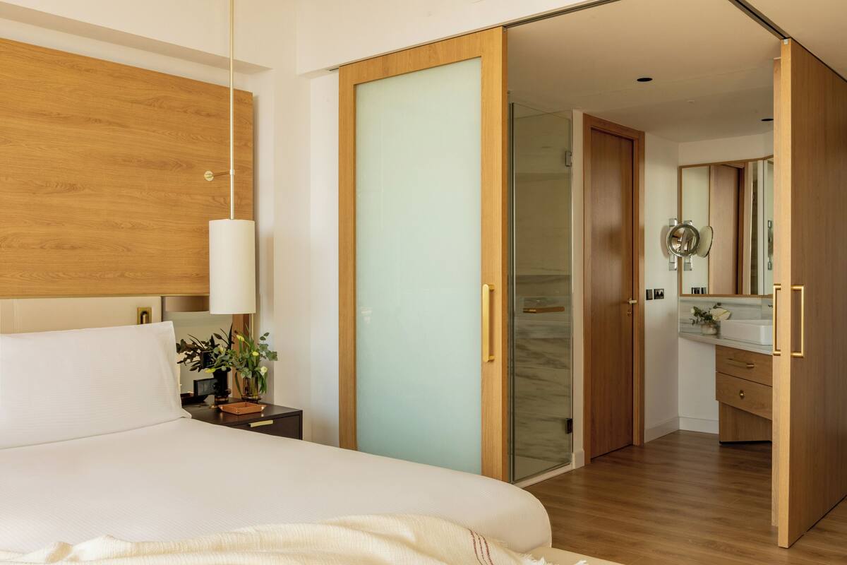 Una habitación de hotel moderna con una cama grande, una mesita de noche con lámpara, una puerta corredera que da al baño y un armario de madera.