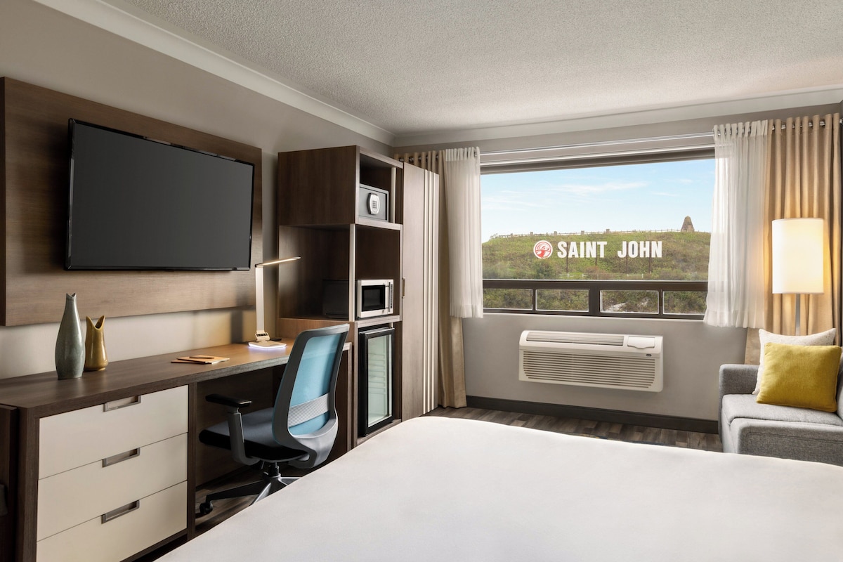 A hotel room with a bed, desk, chair, TV, air conditioning unit, and a window with a view of Saint John.