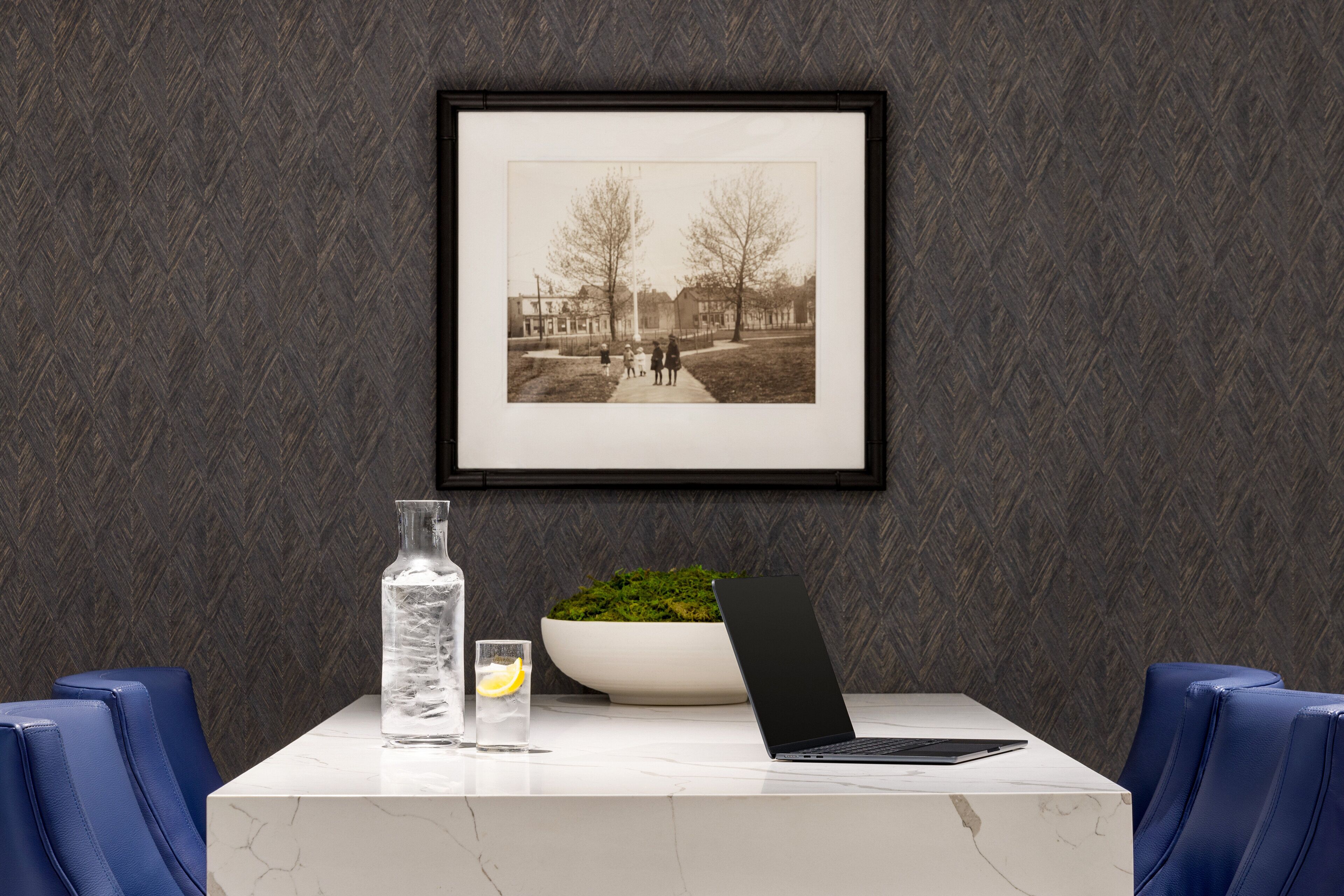 A framed black and white photograph on a wall, a laptop on a marble table, a glass of water, and a bowl of green plants.