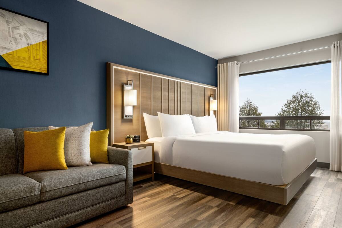 Habitación de hotel con cama, mesita de noche, sofá y un amplio ventanal con vistas.