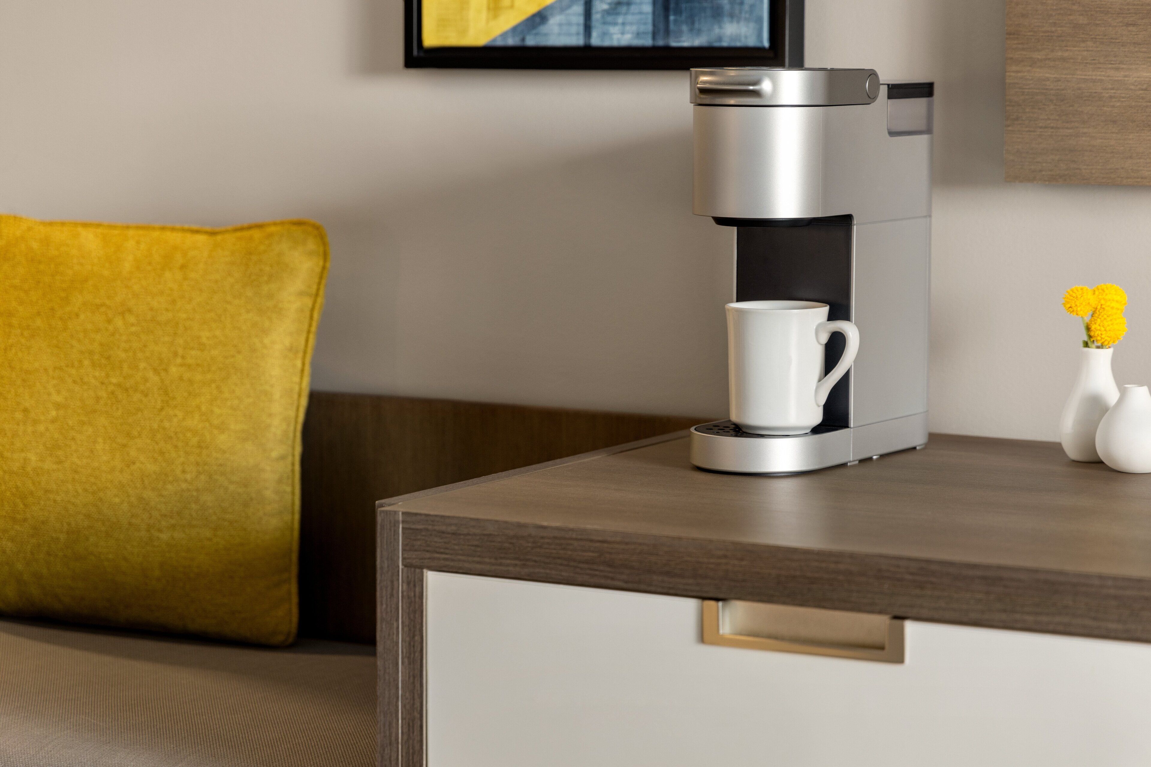 A modern coffee maker on a wooden table with a white mug, a yellow pillow, and a vase with yellow flowers.