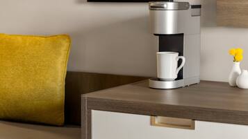 A modern coffee maker on a wooden table with a white mug, a yellow pillow, and a vase with yellow flowers.