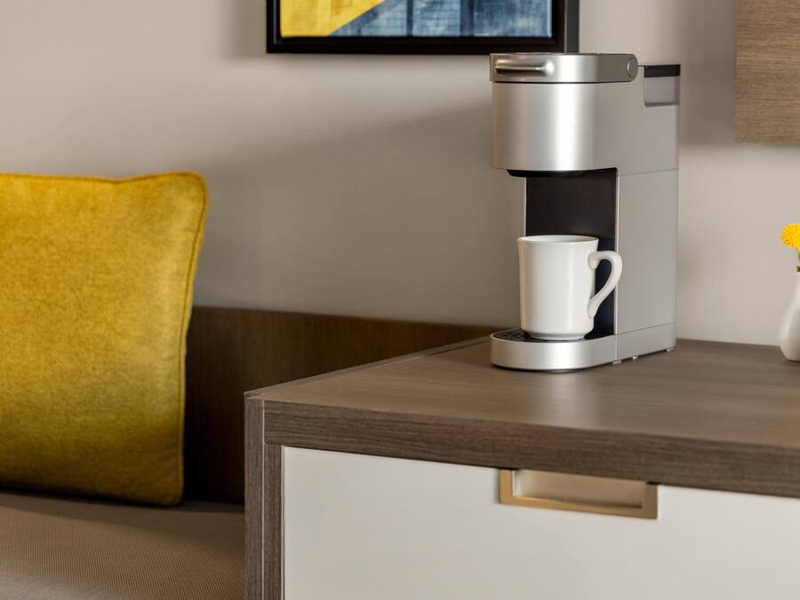 A modern coffee maker on a wooden table with a white mug, a yellow pillow, and a vase with yellow flowers.