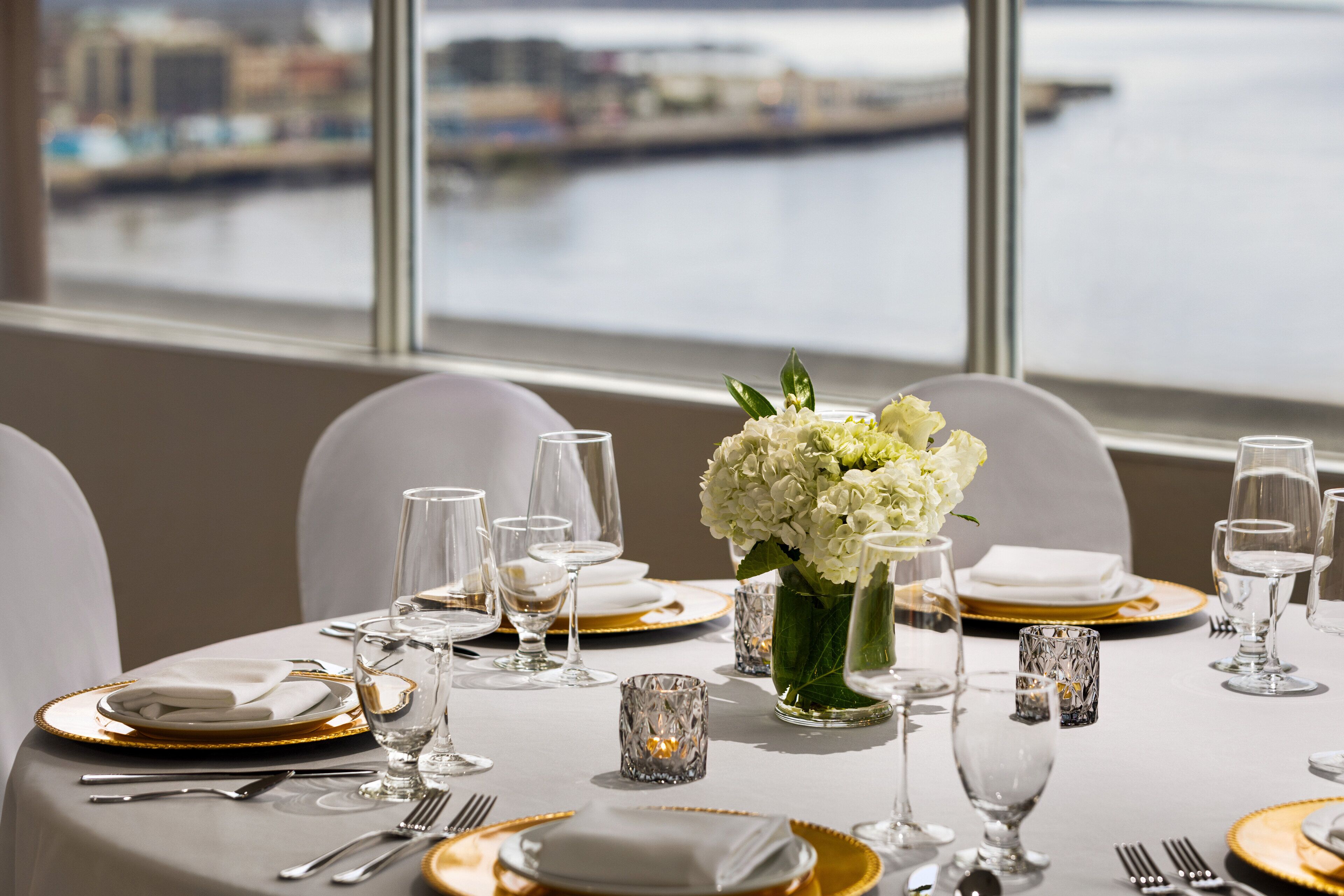 A table set for a formal dinner with white and gold tableware, a vase of white flowers, and a view of a body of water through large windows.