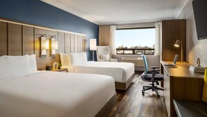 Habitación de hotel con dos camas, un escritorio, una silla, una lámpara y vistas a la ciudad.