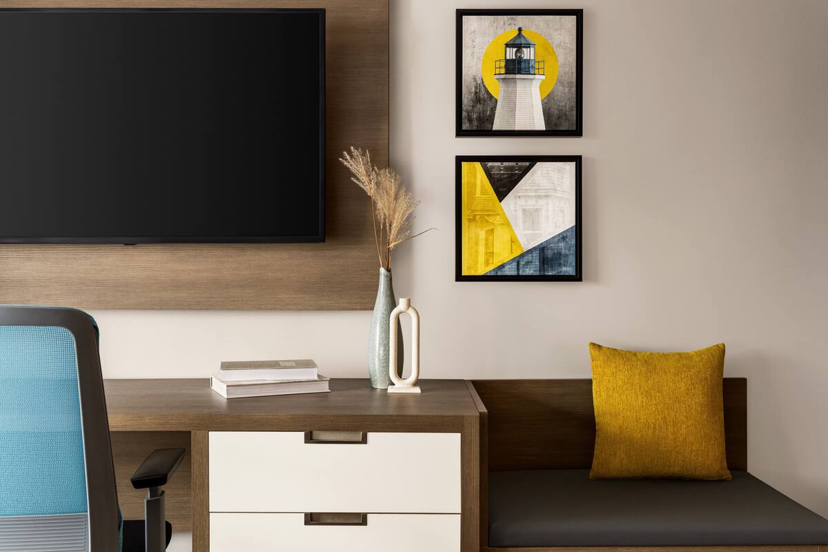 Una habitación moderna con un escritorio de madera, un televisor negro, un sofá con un cojín amarillo y dos obras de arte enmarcadas.