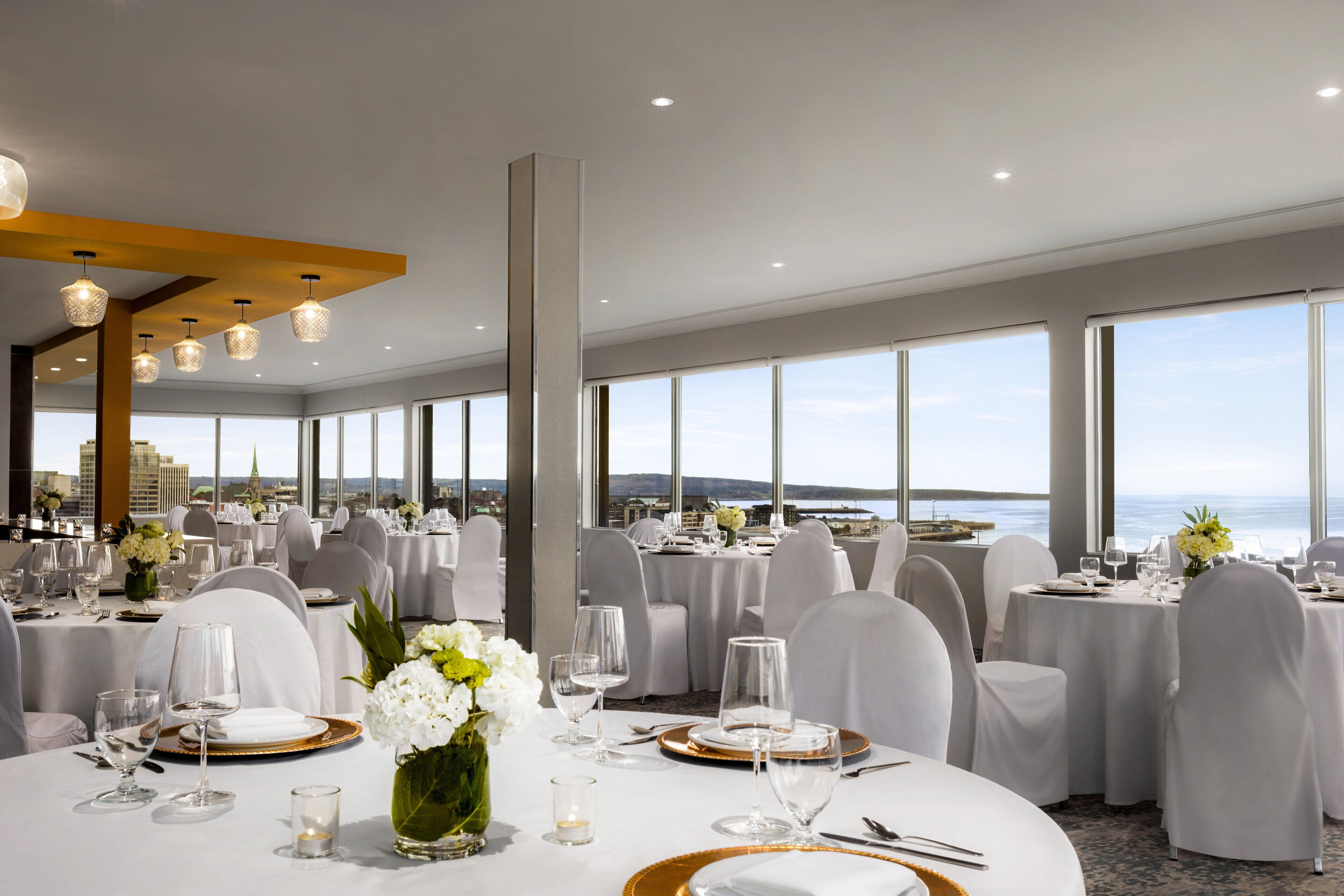 Une salle de banquet avec des tables rondes dressées pour le repas, de grandes fenêtres offrant une vue sur la mer et un design de plafond moderne.