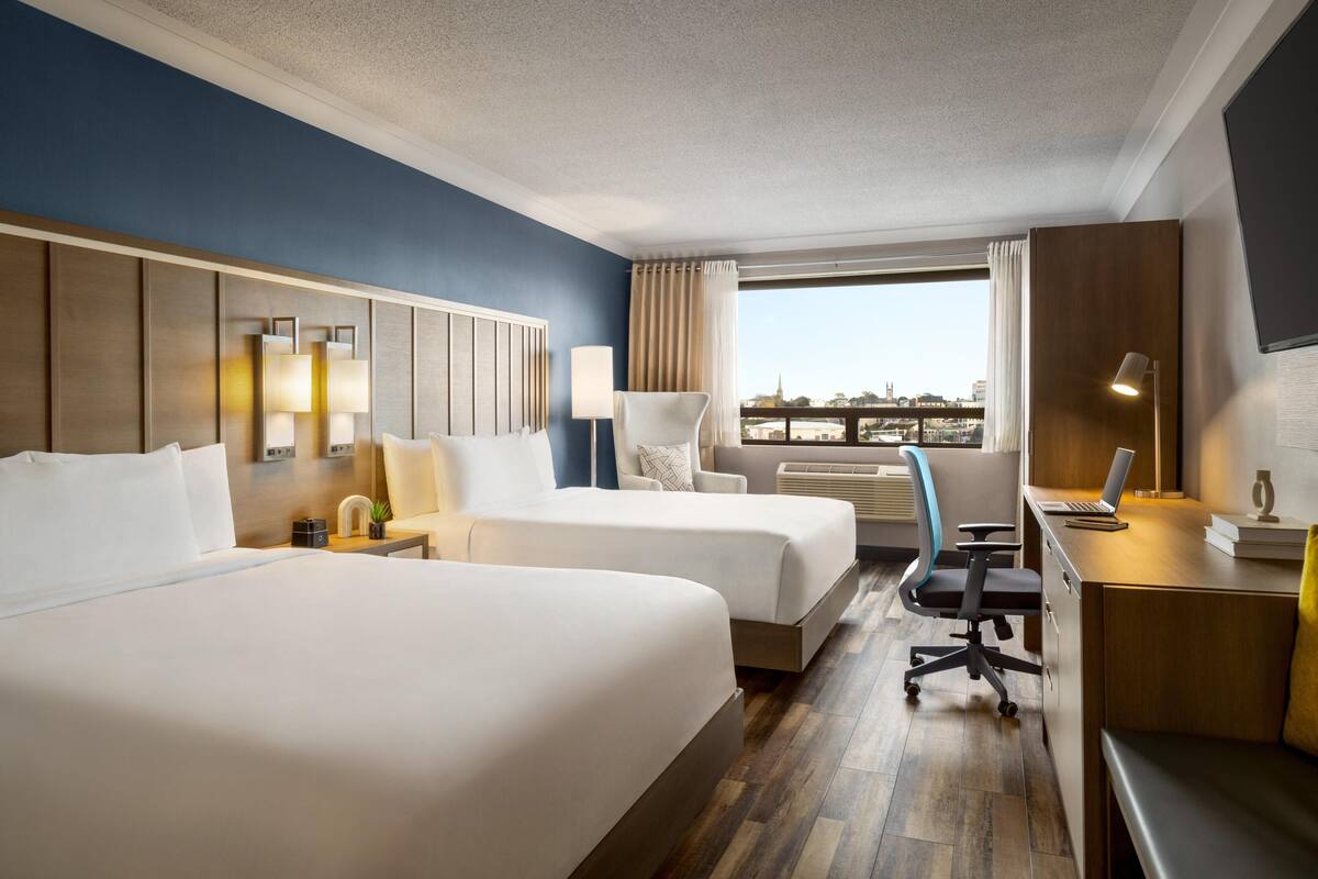 A hotel room with two beds, a desk, a chair, a lamp, and a large window with a city view.