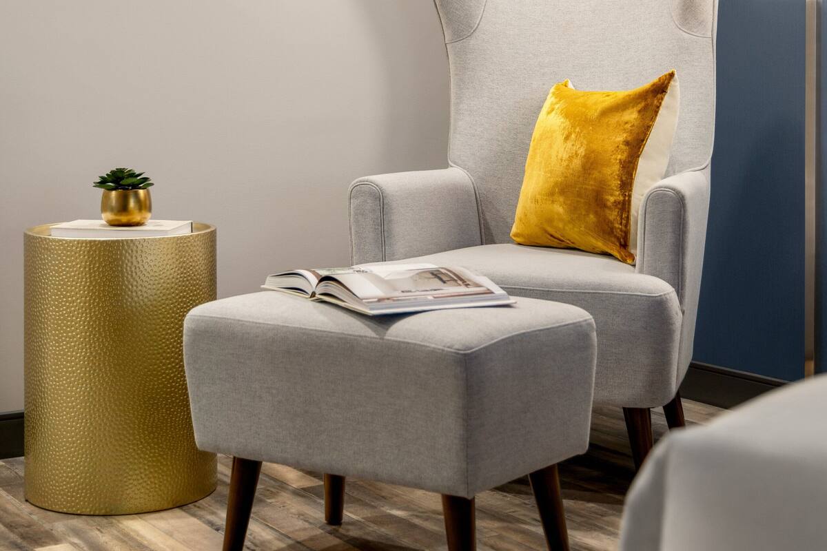 Una habitación moderna con un sillón gris, un cojín amarillo, una pequeña mesa redonda con una planta y una obra de arte enmarcada en la pared.