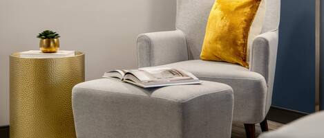 A modern room with a grey armchair, a yellow pillow, a small round table with a plant, and a framed artwork on the wall.