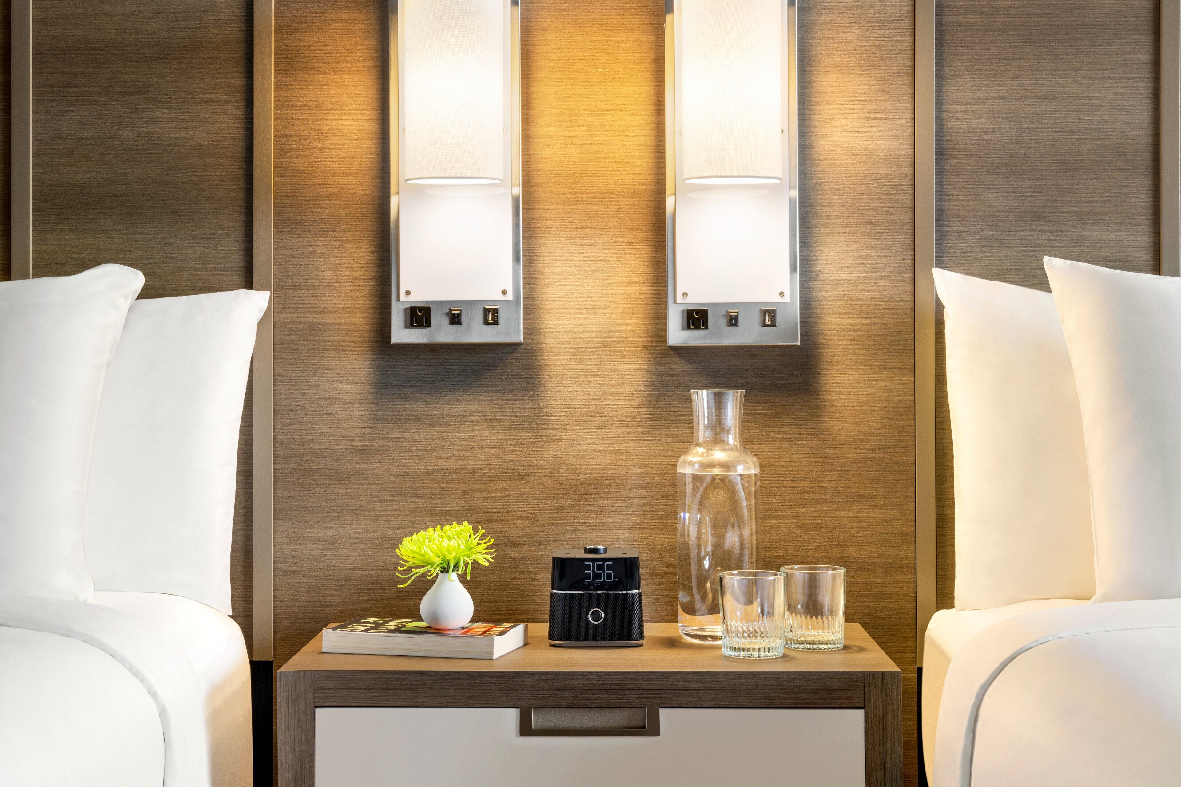 A hotel room with a wooden headboard, two bedside lamps, a nightstand with a digital clock, a vase with a plant, a bottle, and two glasses.