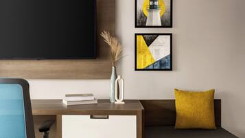 Una habitación moderna con televisión de pantalla plana en la pared, un escritorio con un florero y libros, un sofá con un cojín amarillo, y dos obras de arte enmarcadas.