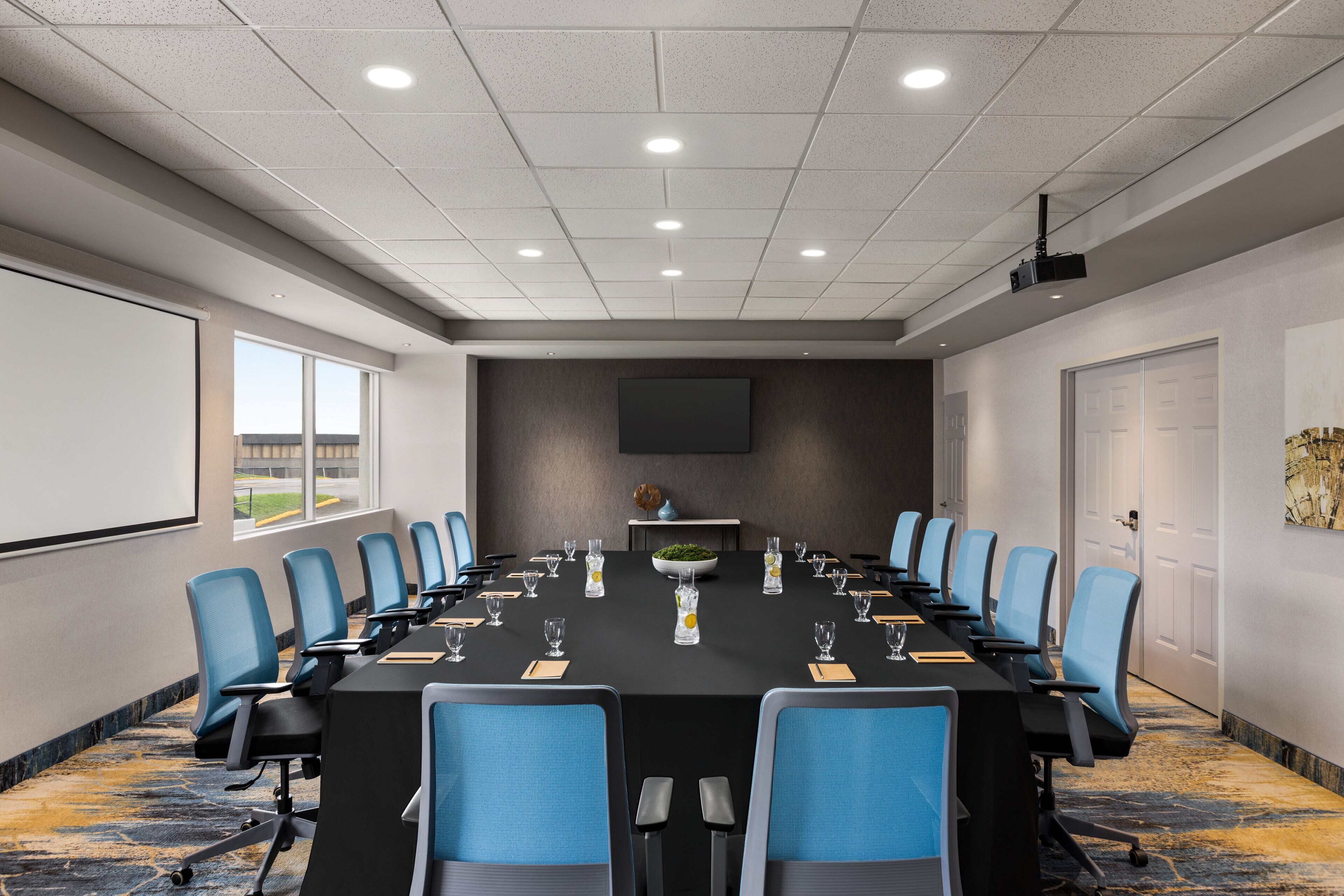 Une salle de conférence avec une longue table, des chaises bleues et un écran de projection.