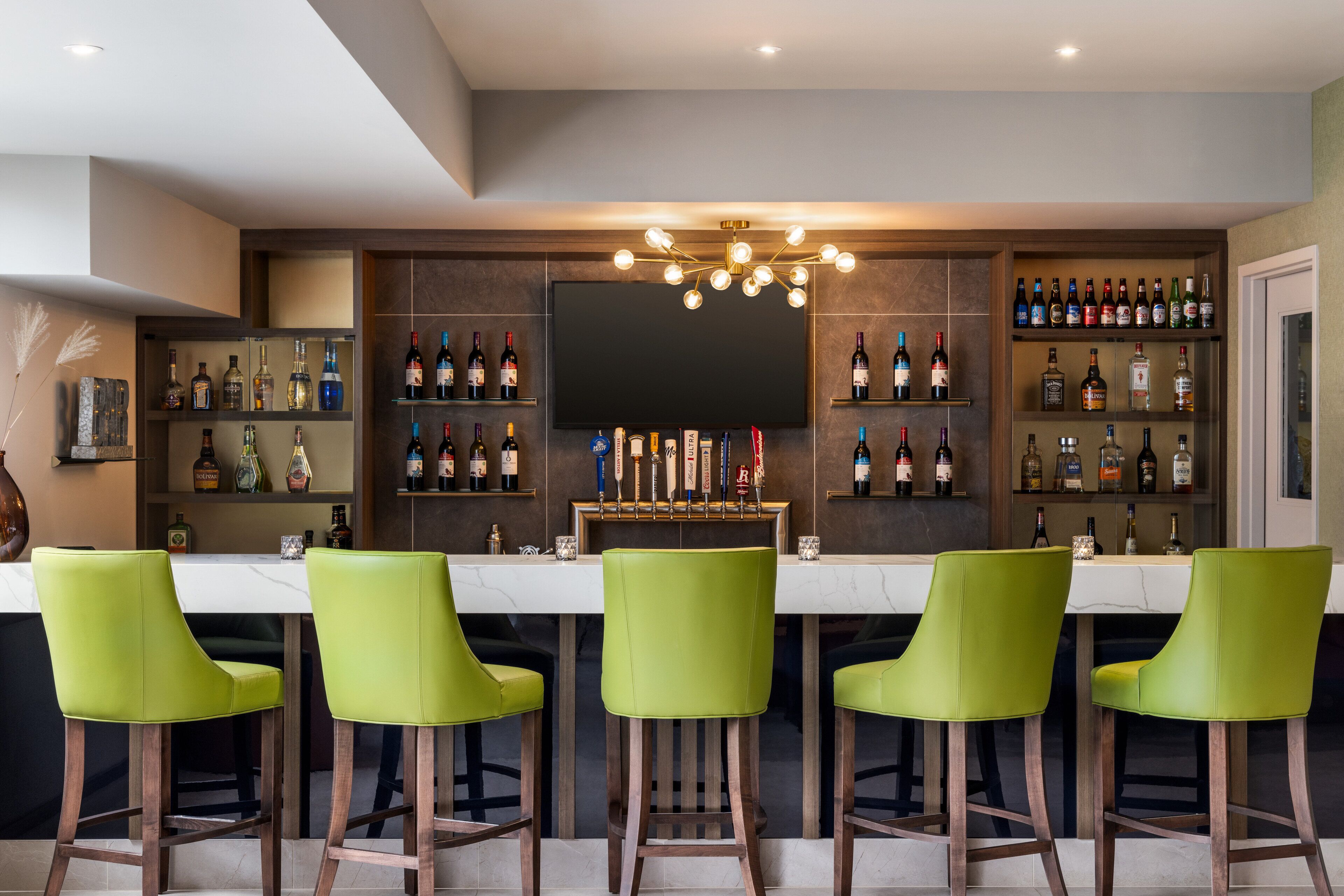 Een bar met een lange toonbank, meerdere planken vol drankflessen en groene barkrukken.