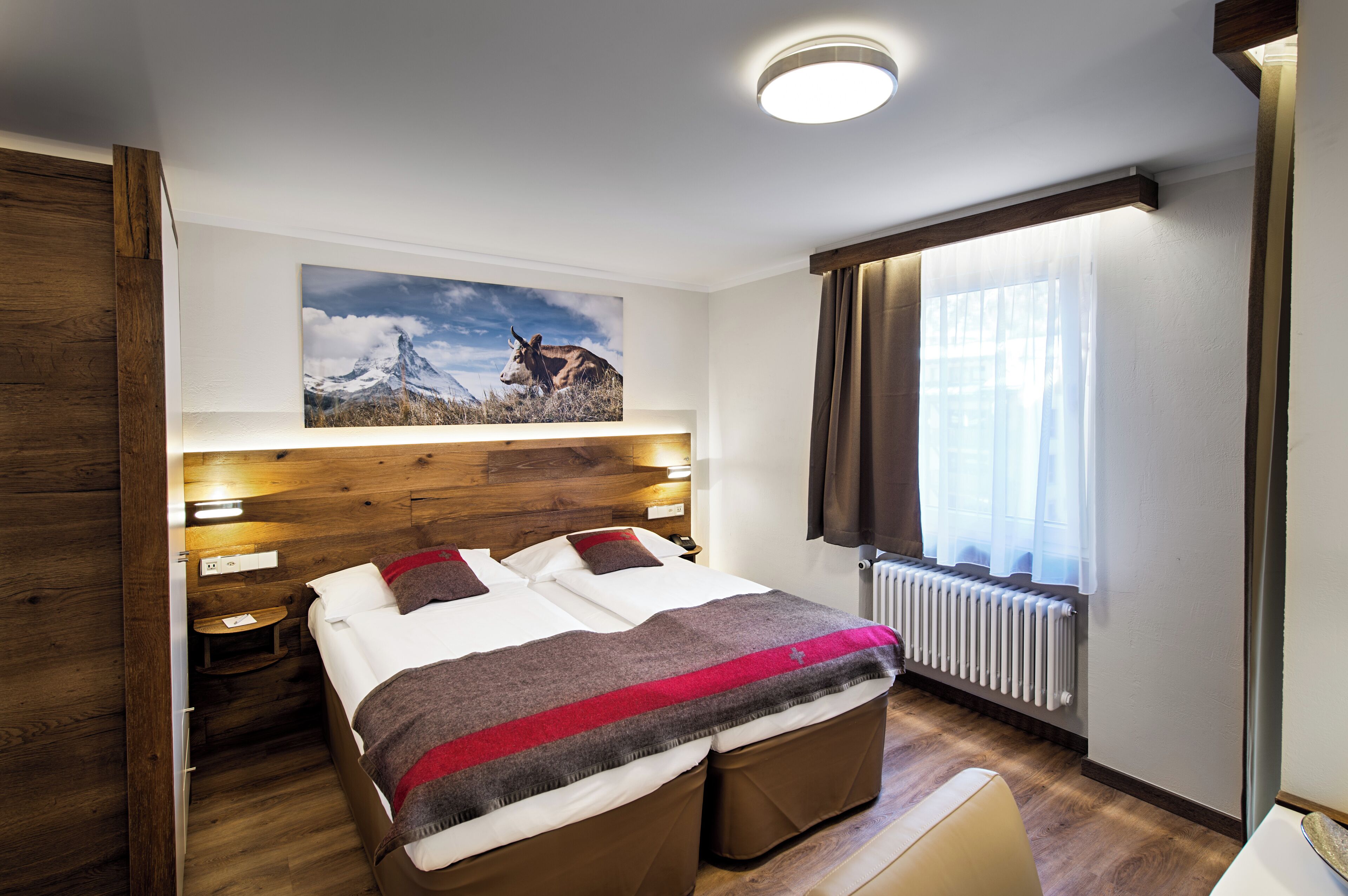 Habitación de hotel moderna con cabecera de madera, una cama grande con manta roja y gris, un cuadro colgado con una montaña y una vaca, una ventana con cortinas y una mesita de noche con lámpara.
