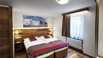 Habitación de hotel moderna con cabecera de madera, una cama grande con manta roja y gris, un cuadro colgado con una montaña y una vaca, una ventana con cortinas y una mesita de noche con lámpara.