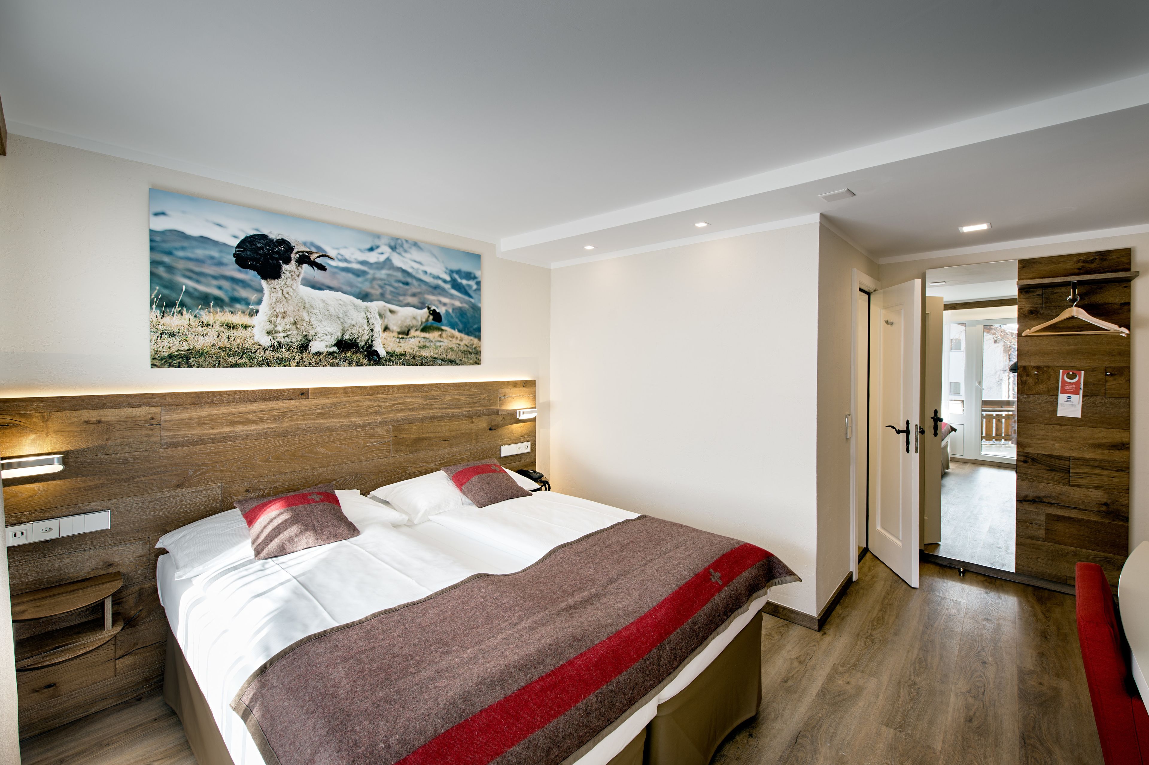A hotel room with a bed, a wooden headboard, a painting of sheep on the wall, and a wooden wardrobe.