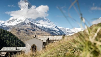 A small white building with a sloped roof, surrounded by tall grass and mountains in the background.