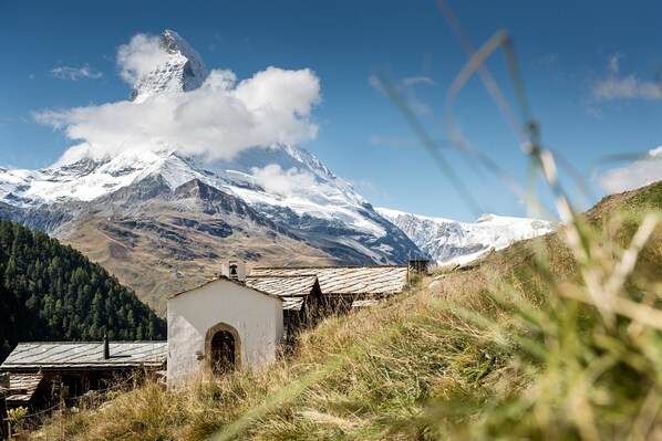 A small white building with a sloped roof, surrounded by tall grass and mountains in the background.