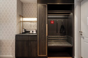 A hotel room with a dark wood vanity, a mirror, a small table with a kettle and cups, and a red sign on the door.