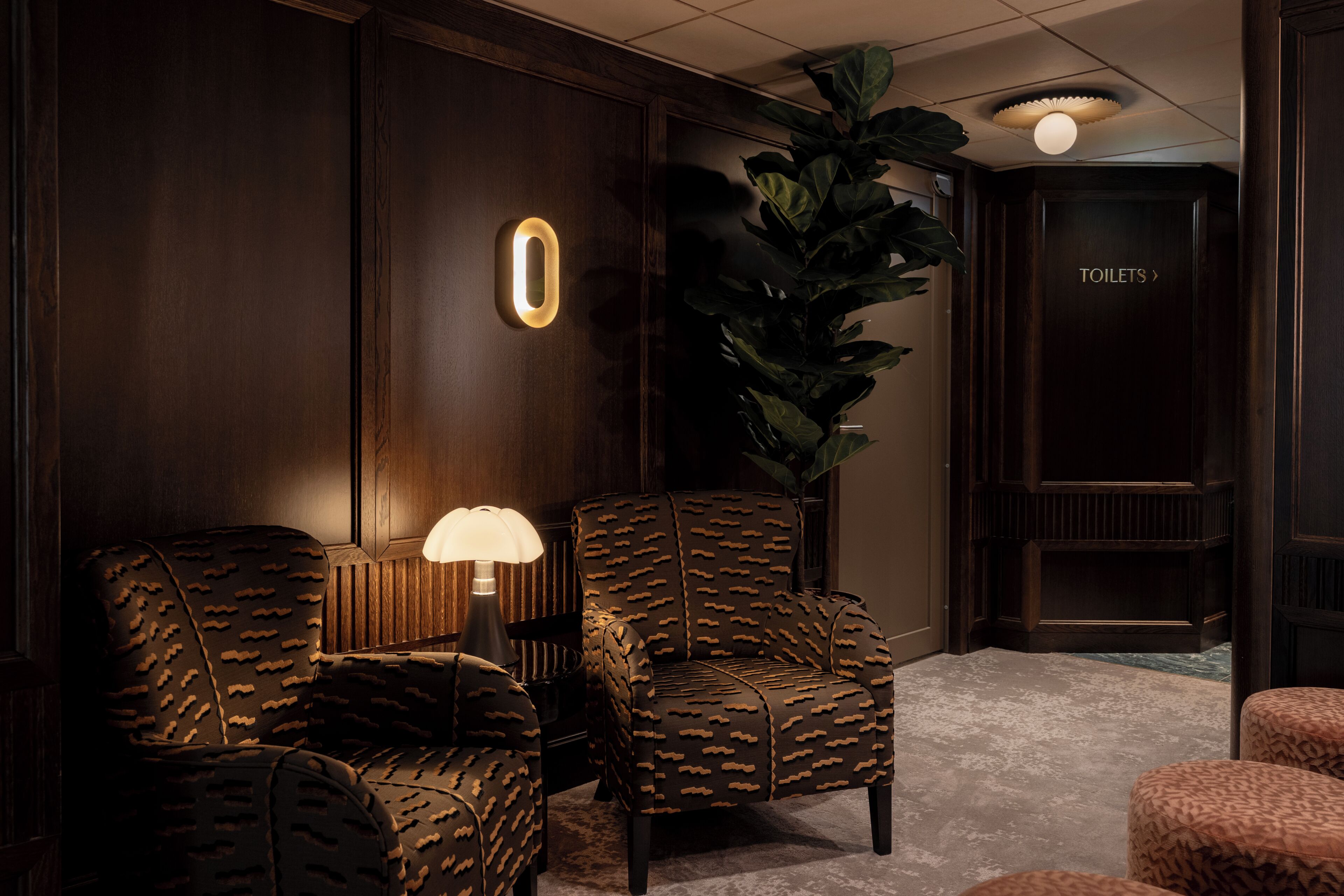 Una habitación revestida de madera oscura con dos sillones estampados, una lámpara de pie y una planta grande en maceta. Se ve una puerta con la inscripción 'TOILETS'.