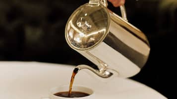 En hånd hælder kaffe fra en metalkande i en hvid kop på en underkop.