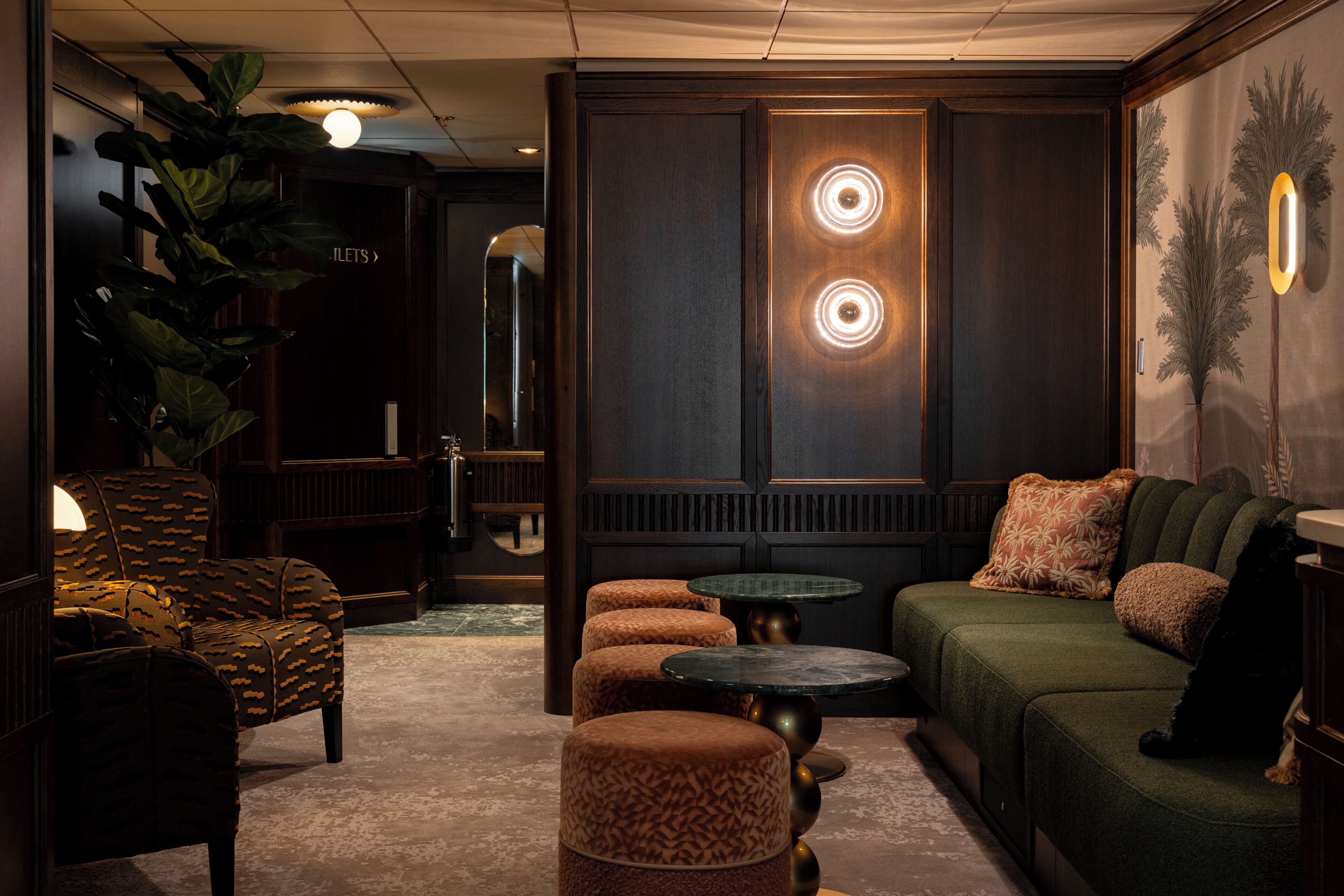 Un salon faiblement éclairé, avec un canapé, des poufs, une table ronde et une grande plante.