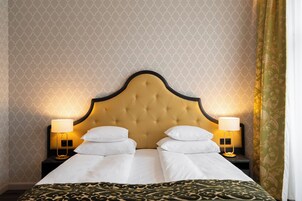 Uma cama de casal com cabeceira estofada, duas mesas de cabeceira com luminárias e um edredom estampado.
