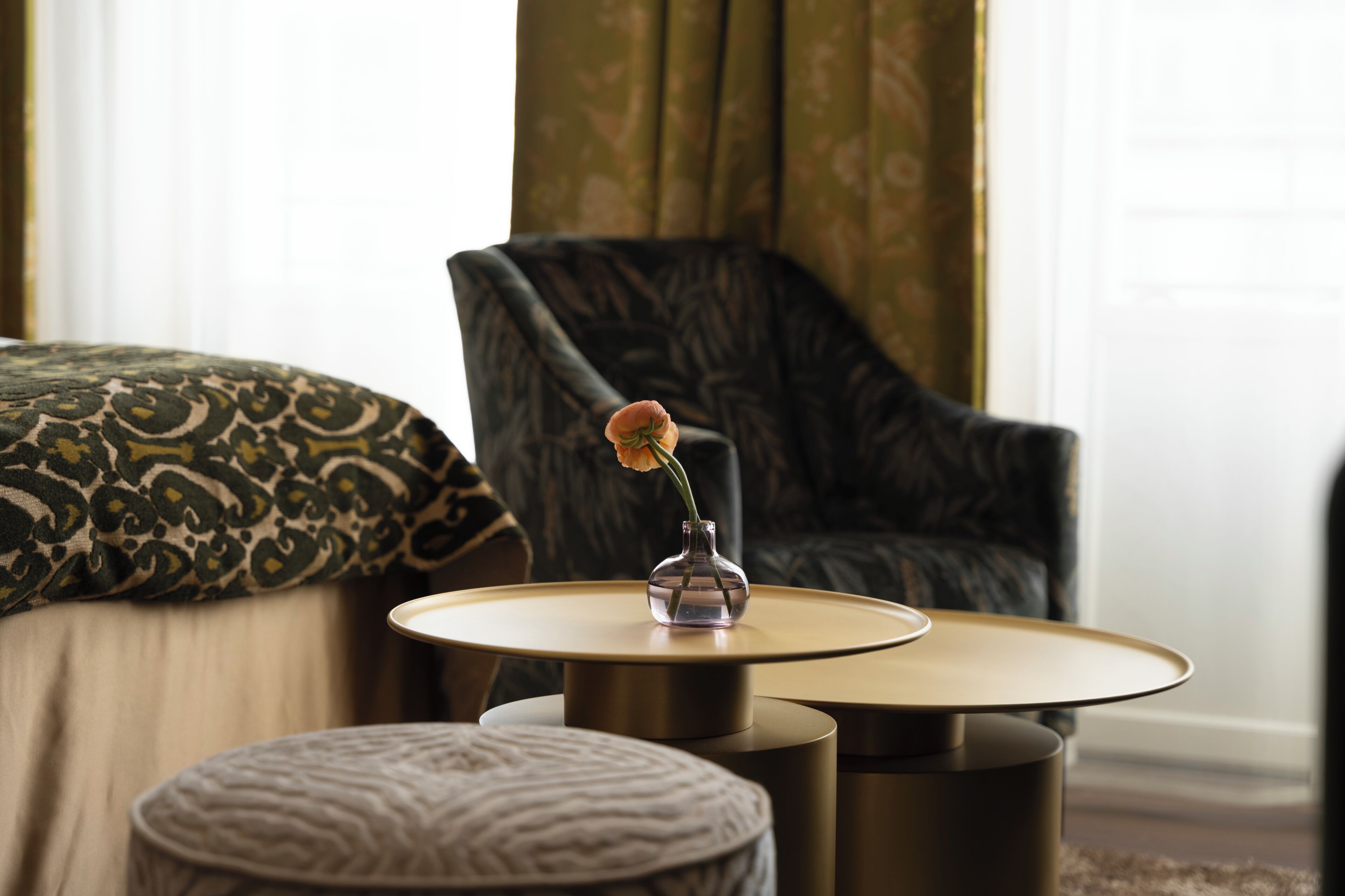 A room with a bed, a chair, a small table with a vase, and a patterned pillow.