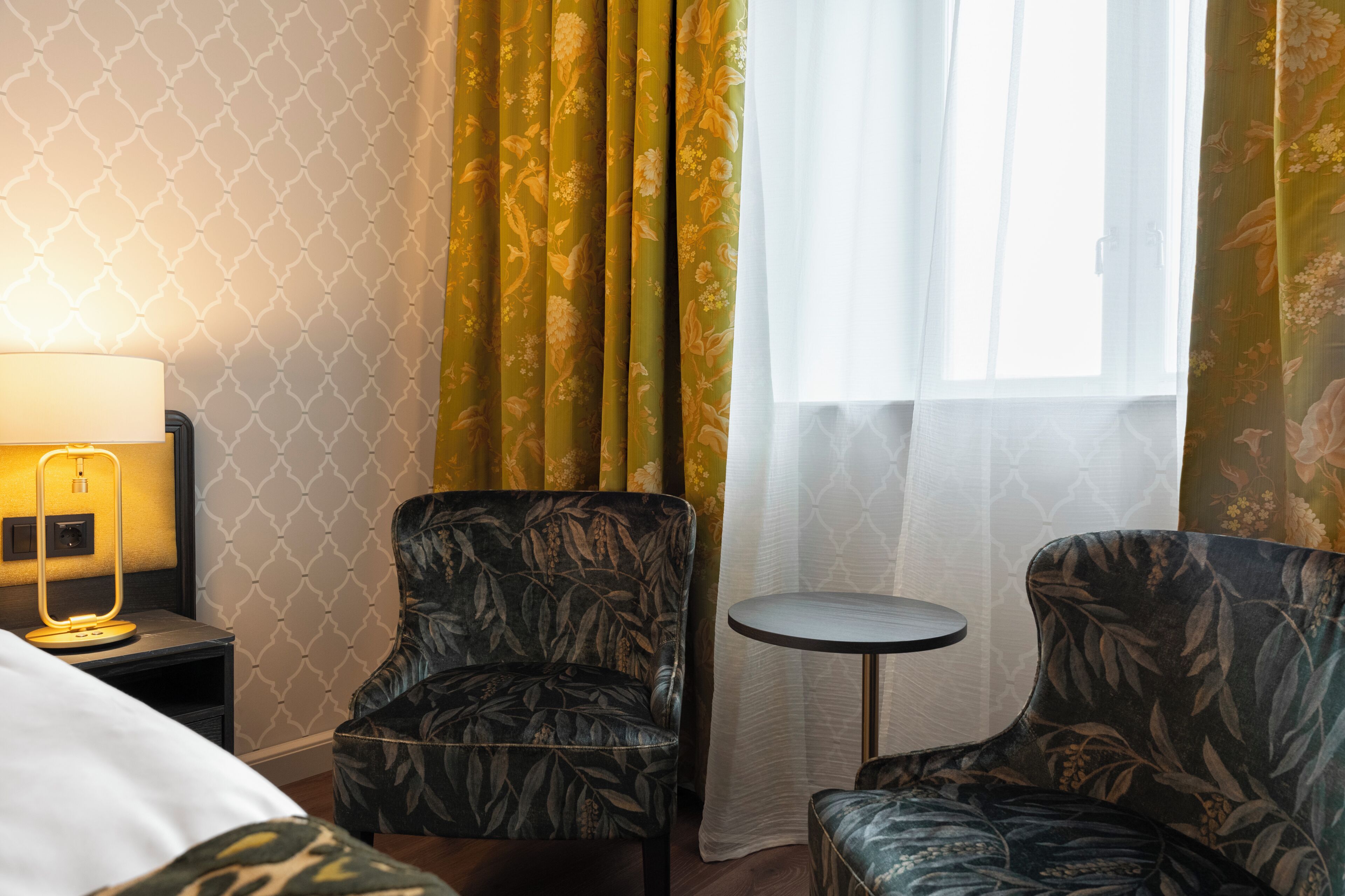 A hotel room with a bed, two armchairs, a small round table, a lamp, and a window with curtains.