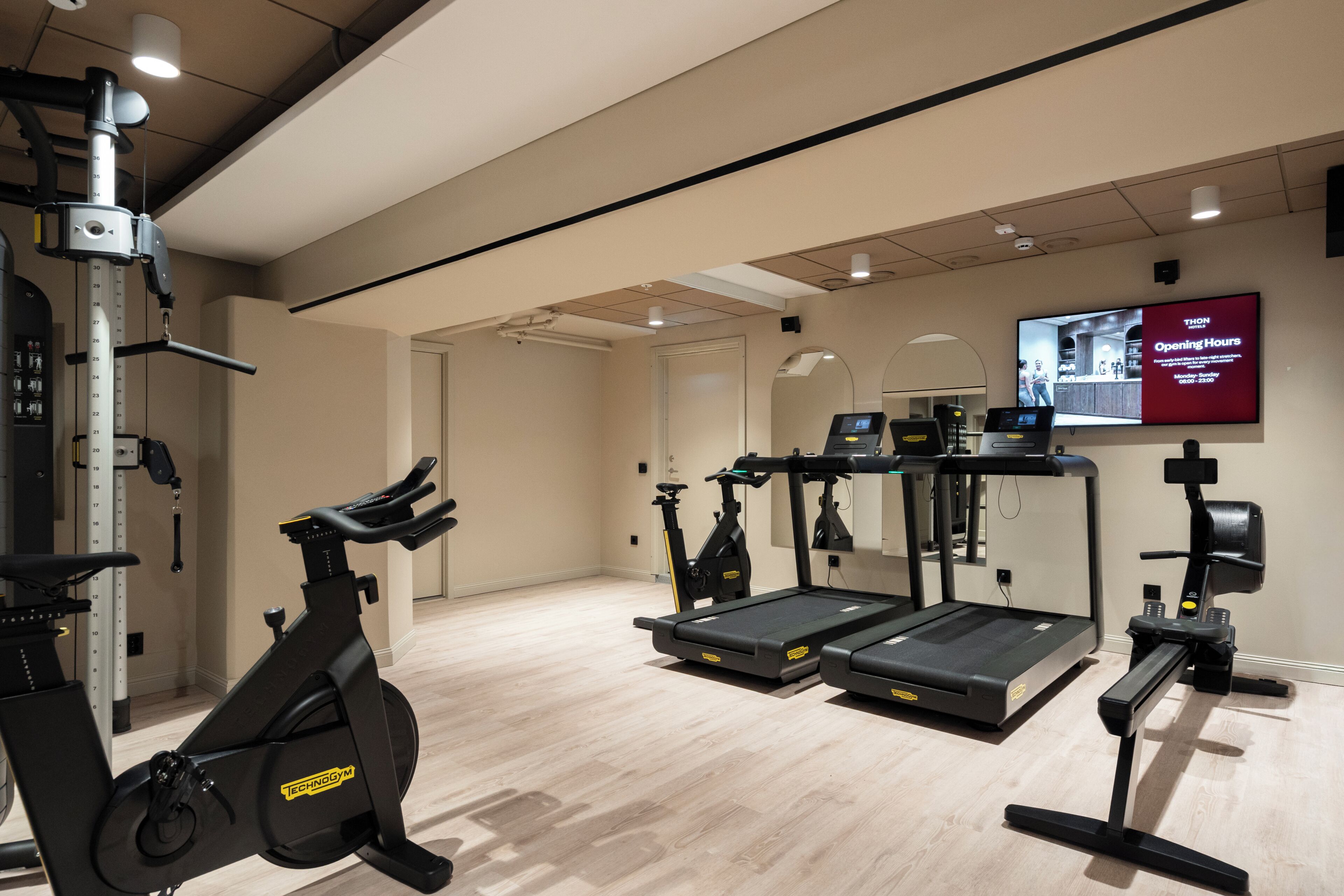 Une salle de sport équipée de matériel d’exercice, un téléviseur affichant les heures d’ouverture et une porte donnant sur une autre pièce.