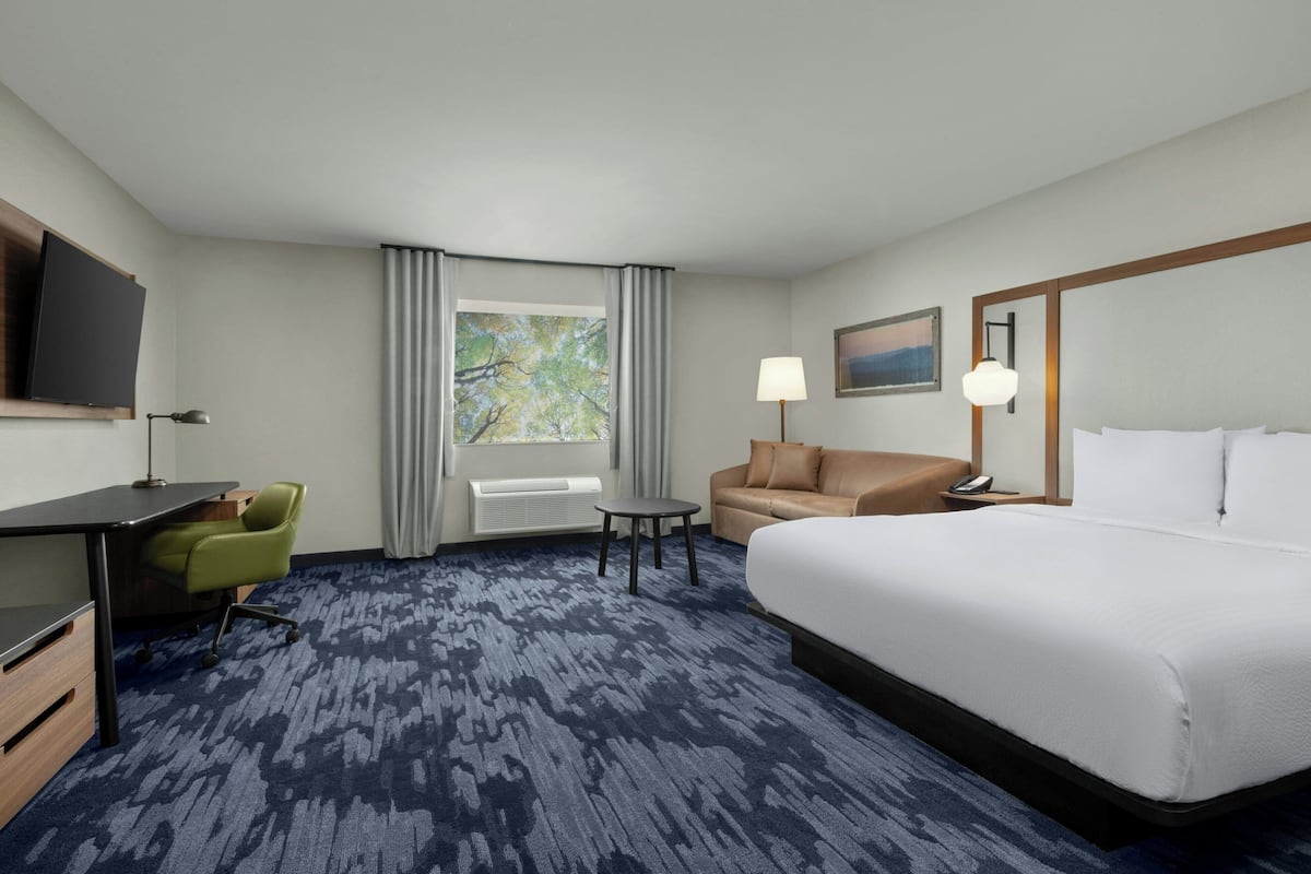 Une chambre d’hôtel avec un grand lit, un bureau, une chaise, un canapé, une petite table, une lampe, une télévision et une fenêtre avec des rideaux.
