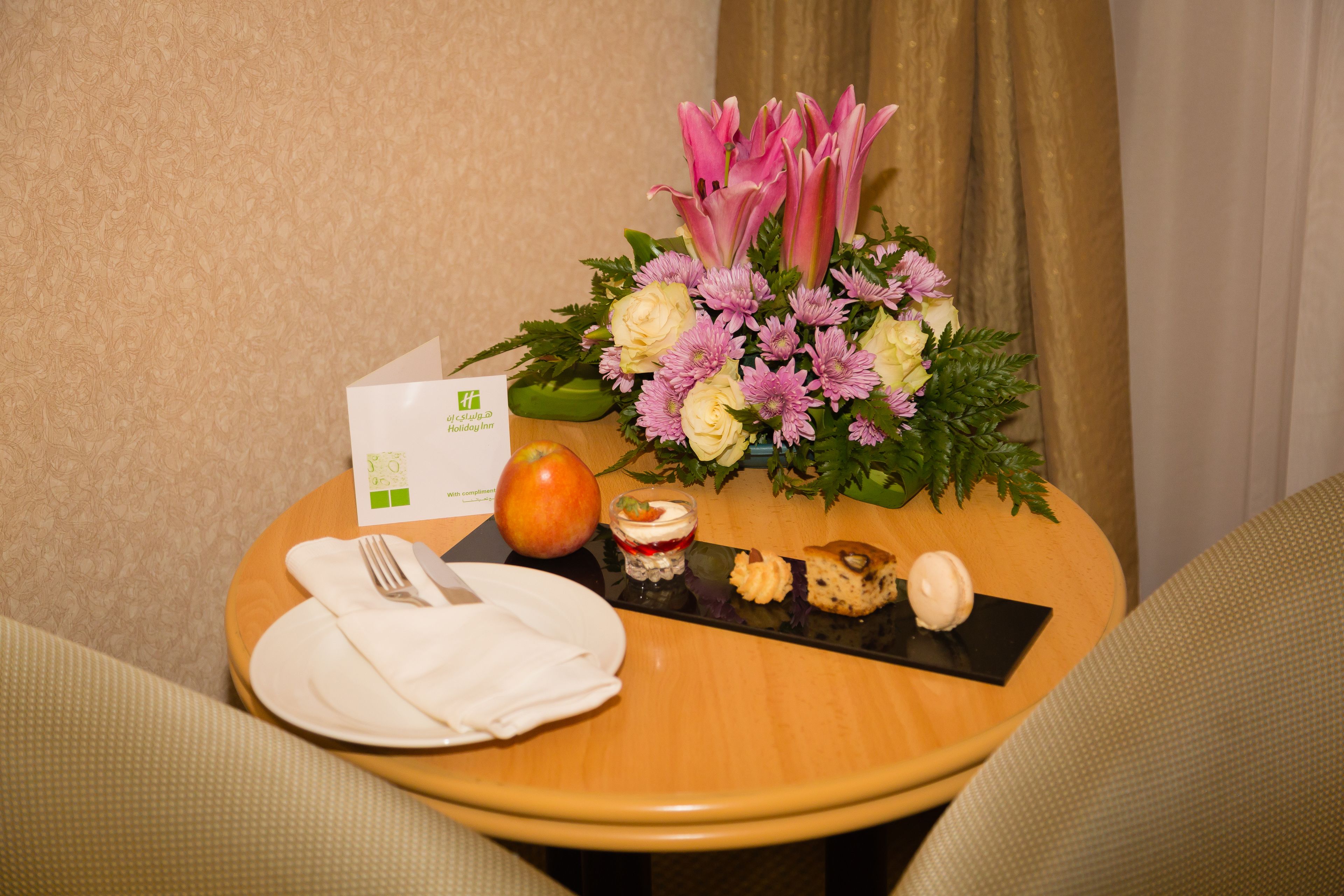 A table with a floral arrangement, a plate with a fork and knife, an apple, a dessert, and a small card.