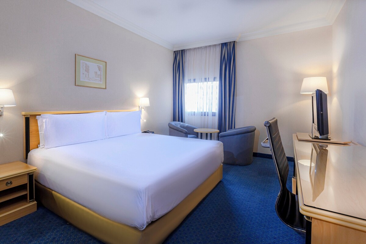 Une chambre d’hôtel comprenant un lit, des tables de chevet, une chaise, un bureau avec une lampe et un téléviseur.