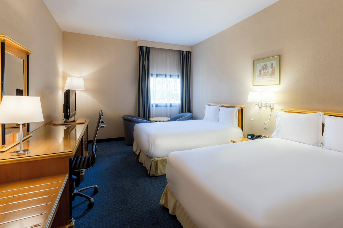 Une chambre d’hôtel avec deux lits, un bureau avec une lampe, une chaise, une lampe de bureau, un téléphone et une fenêtre avec des rideaux.