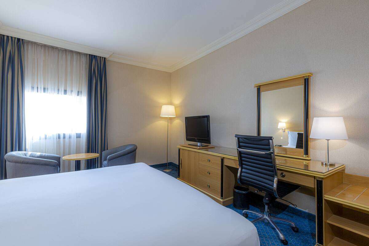 A hotel room with a bed, a desk with a computer, a chair, a TV, and two lamps.