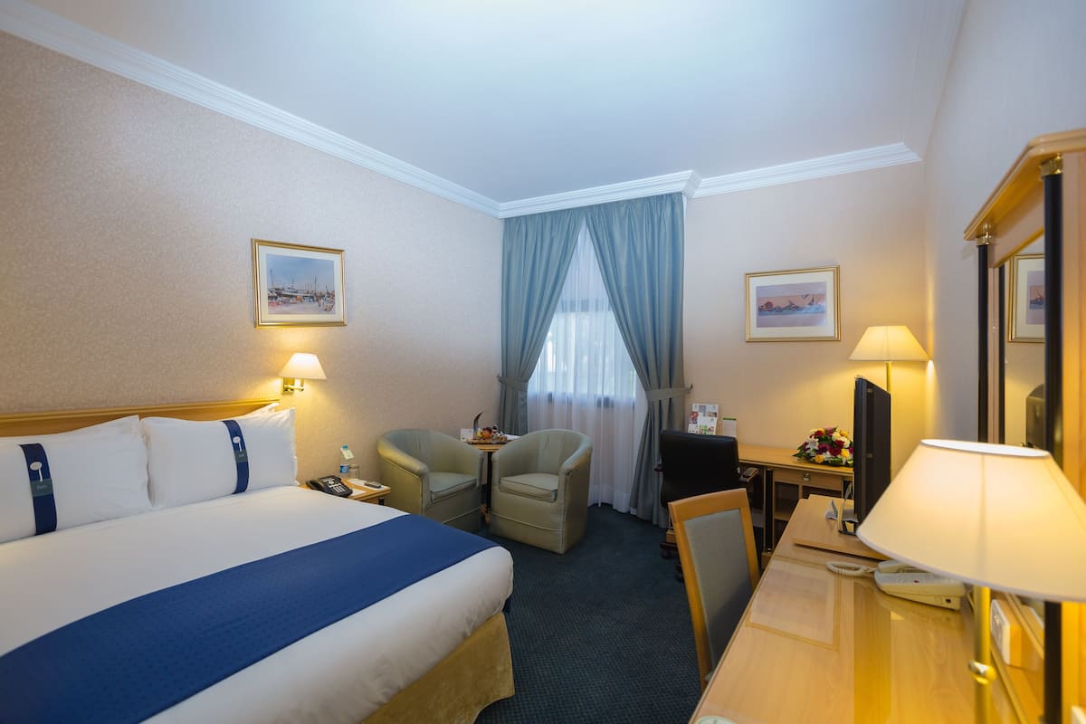 A hotel room with a large bed, two armchairs, a desk with a lamp, and a television.