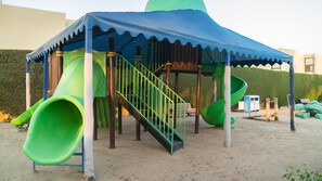 Une aire de jeux pour enfants avec un toboggan et une zone d’escalade, entourée de sable et de verdure.