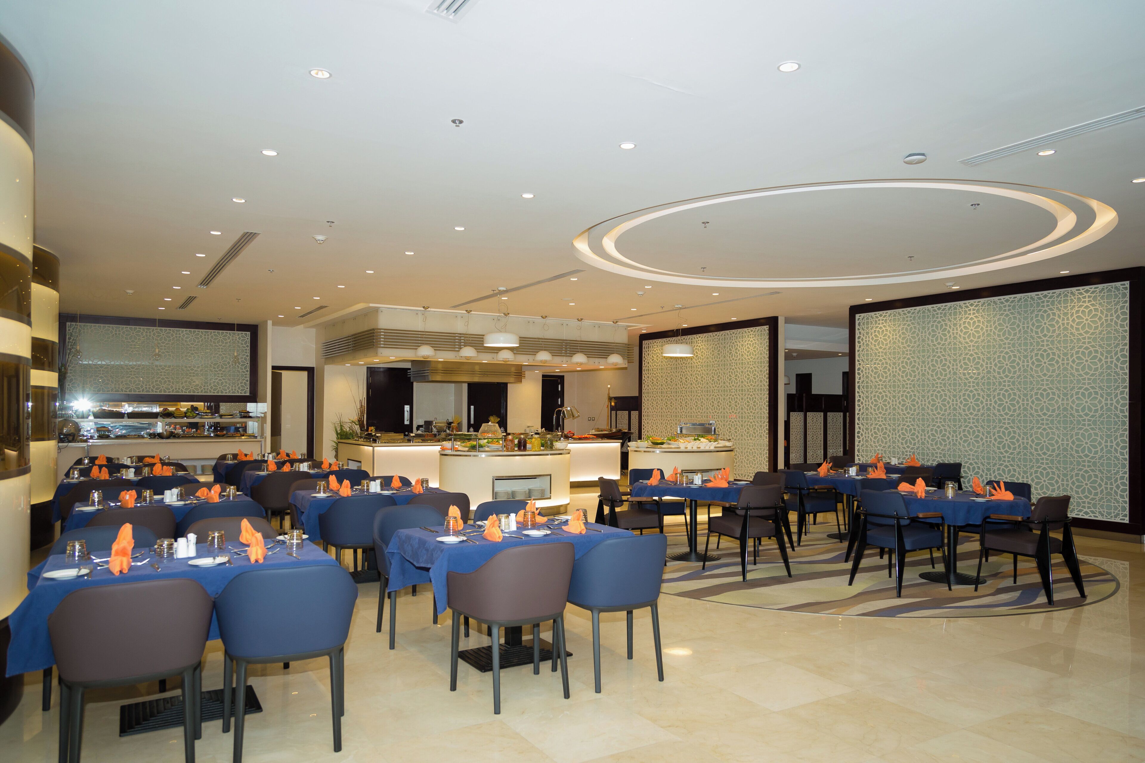 A modern dining area with tables set for service, a kitchen in the background, and a decorative ceiling.