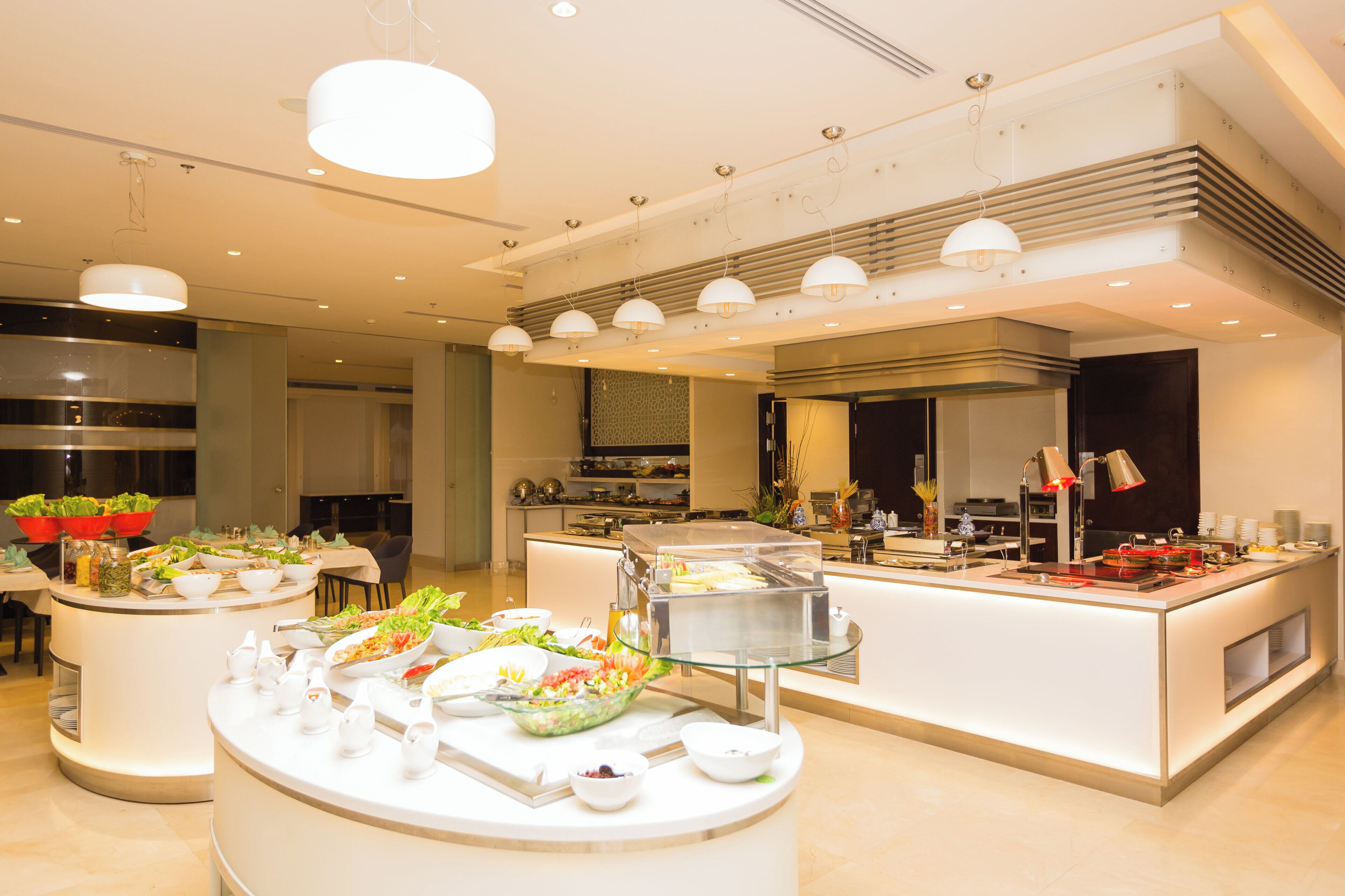 Un comedor moderno de hotel con estaciones de comida tipo buffet, mesas redondas e iluminación contemporánea.