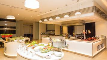 Un comedor moderno de hotel con estaciones de comida tipo buffet, mesas redondas e iluminación contemporánea.