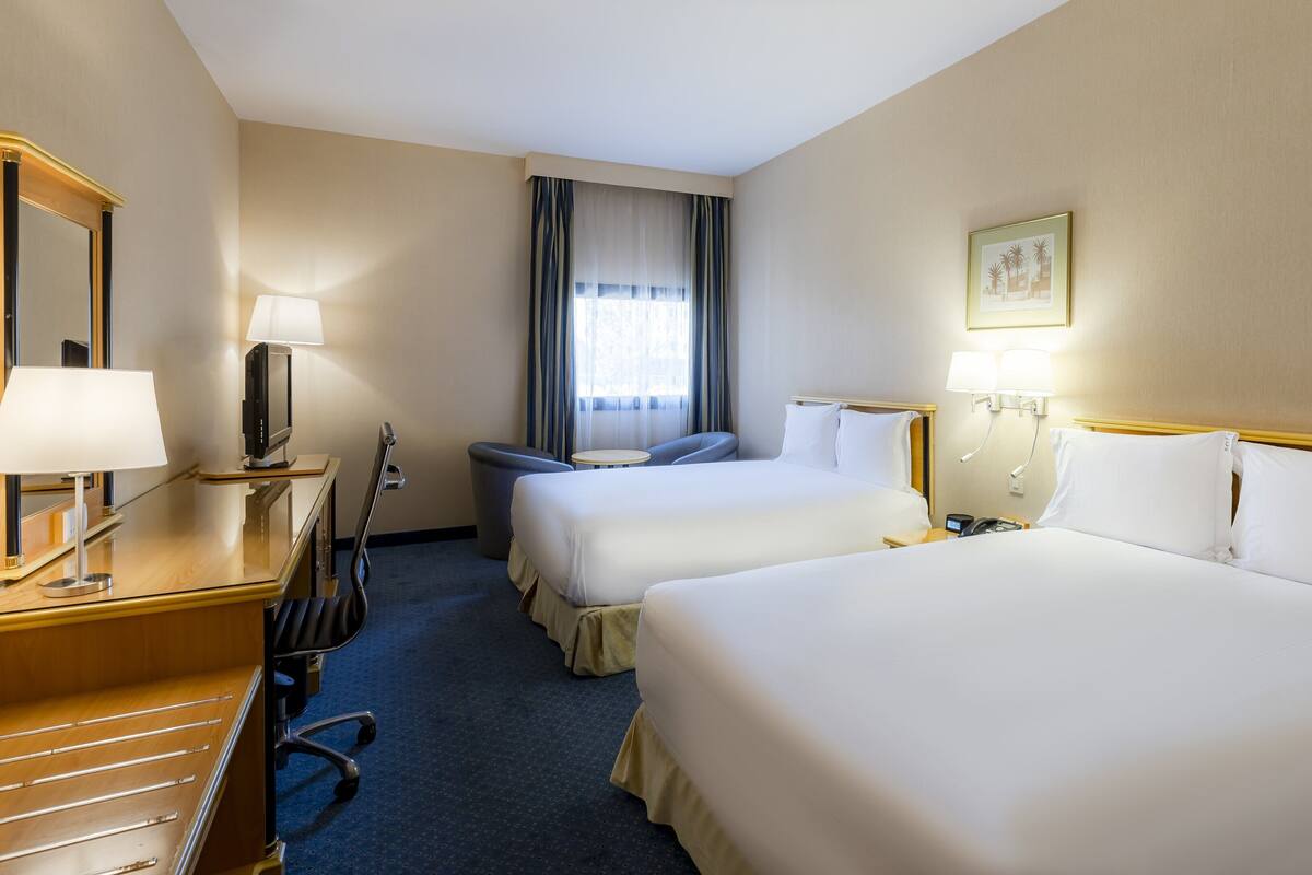 A hotel room with two beds, a desk with a lamp, a chair, a small table, a mirror, and a window with curtains.