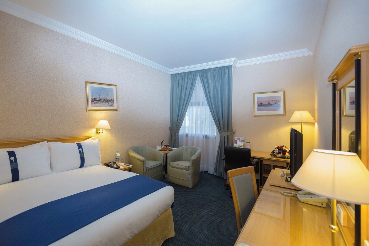 Une chambre d’hôtel comprenant un lit, deux fauteuils, un bureau avec une lampe, une télévision et une fenêtre avec des rideaux.
