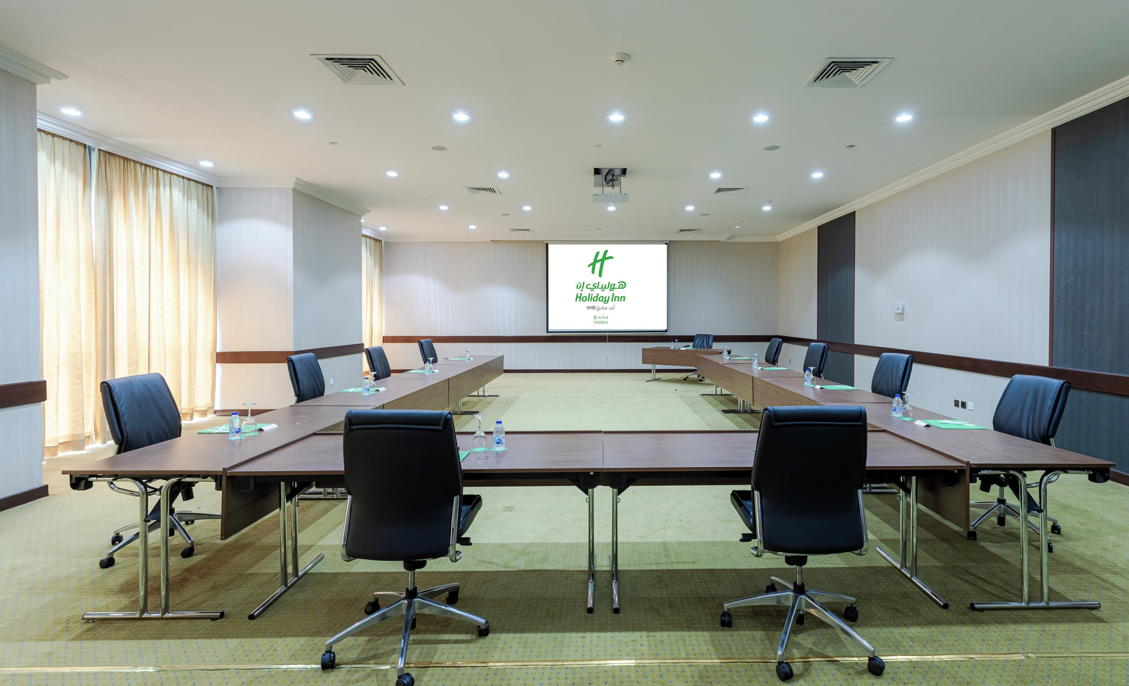 A conference room with a long table, chairs, a projector screen, and a carpeted floor.