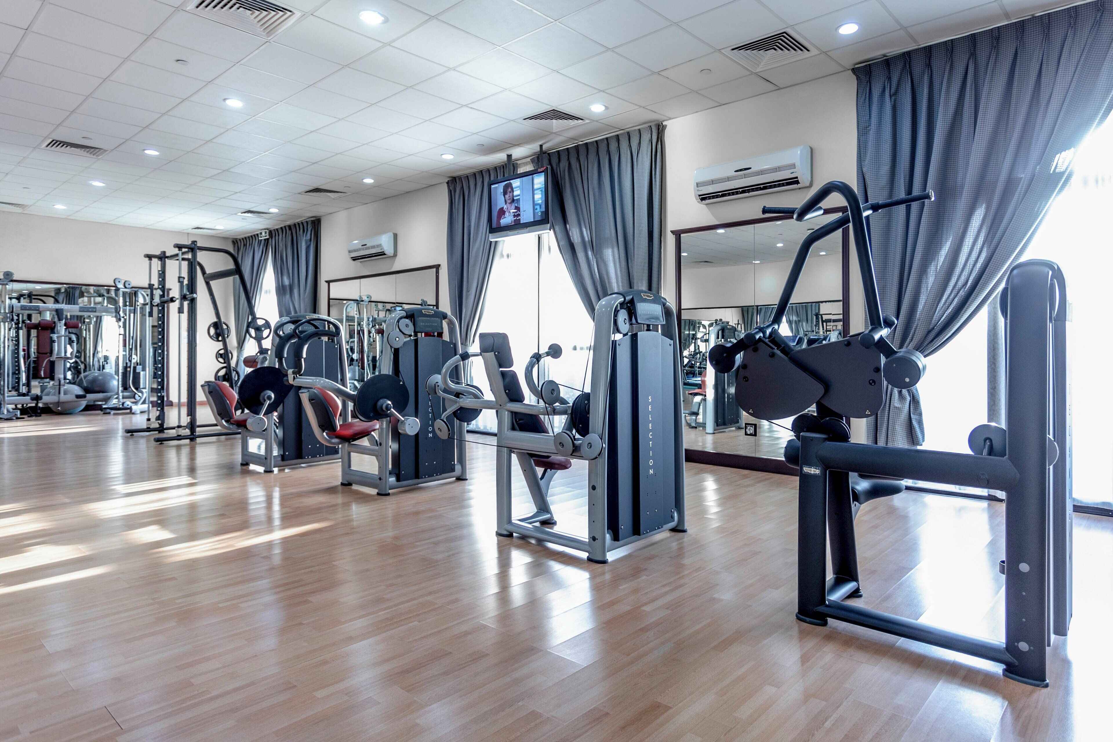 A gym with various exercise machines, a television screen, and large windows with curtains.